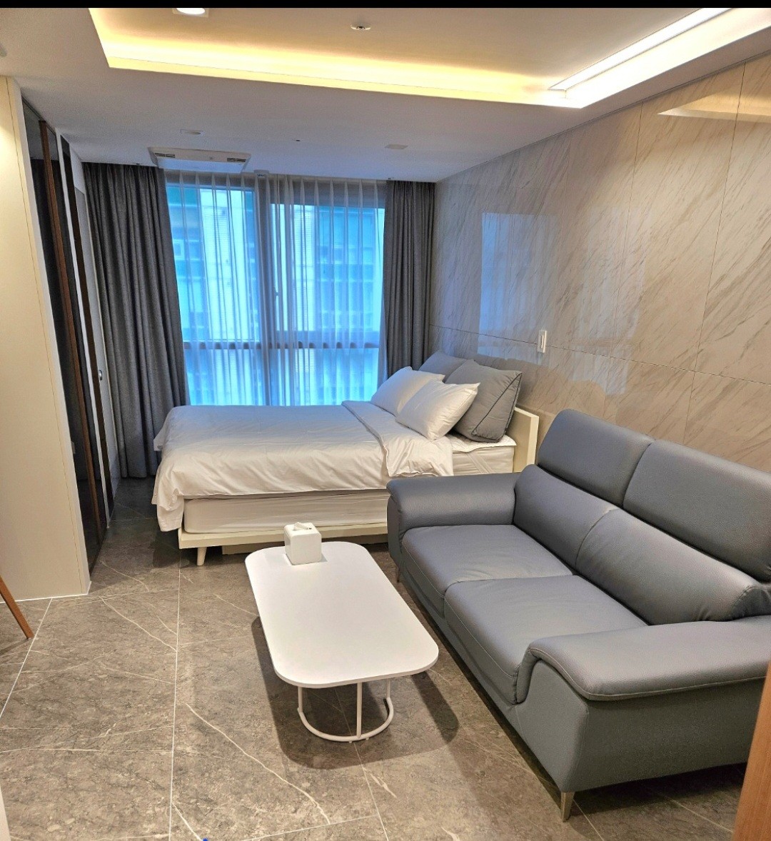 A well-arranged room featuring a spacious bed neatly made with soft linens. A comfortable gray sofa is positioned near a small white coffee table. Large windows are adorned with light curtains, allowing natural light to fill the area. The marble wall adds a touch of elegance.