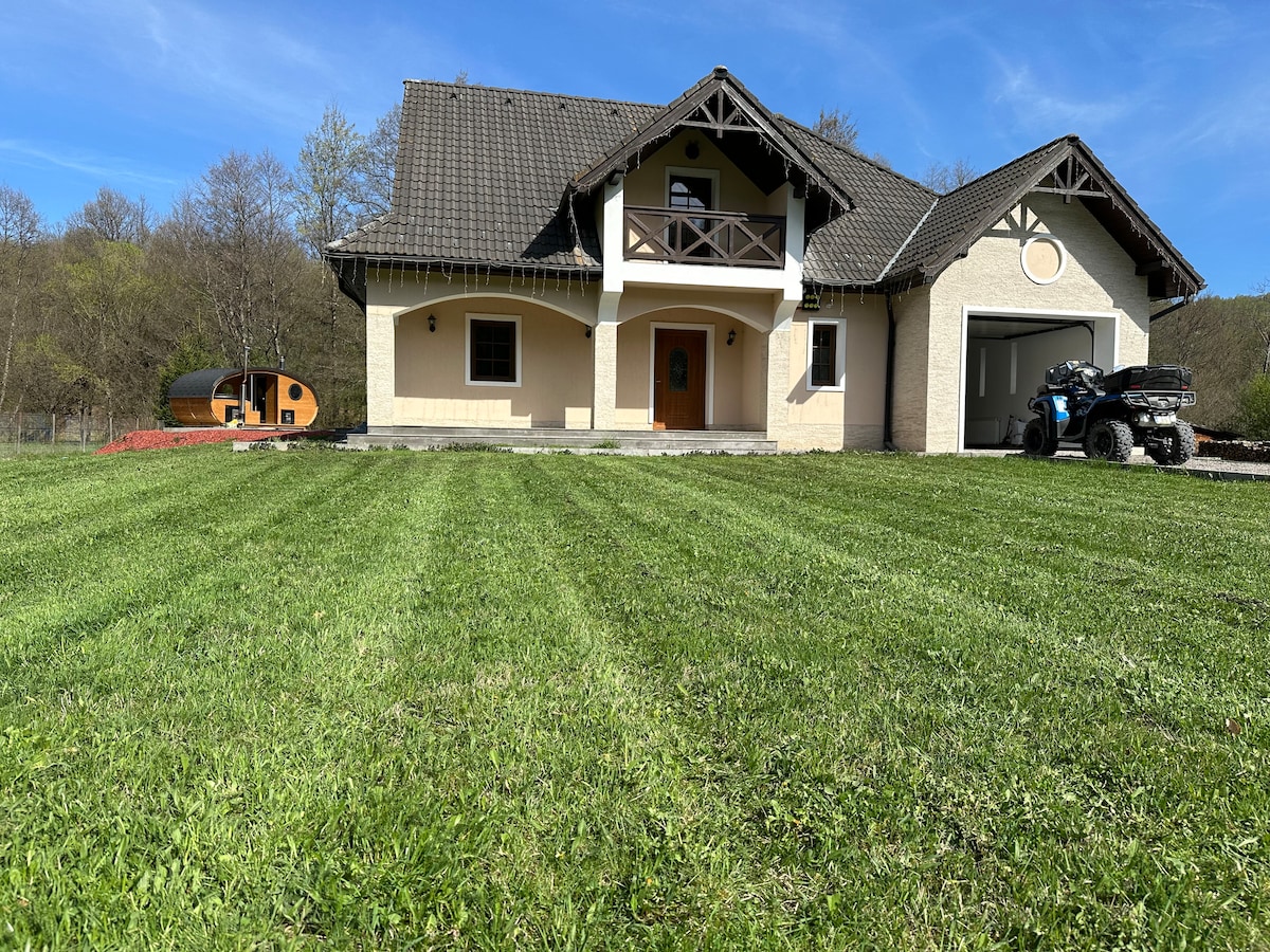 A large, two-story house features a covered front porch and a harmonious blend of white and beige exterior walls. A well-maintained lawn with neatly trimmed grass surrounds the home, complimented by trees in the background. A small cabin and vehicles are visible beside the house.