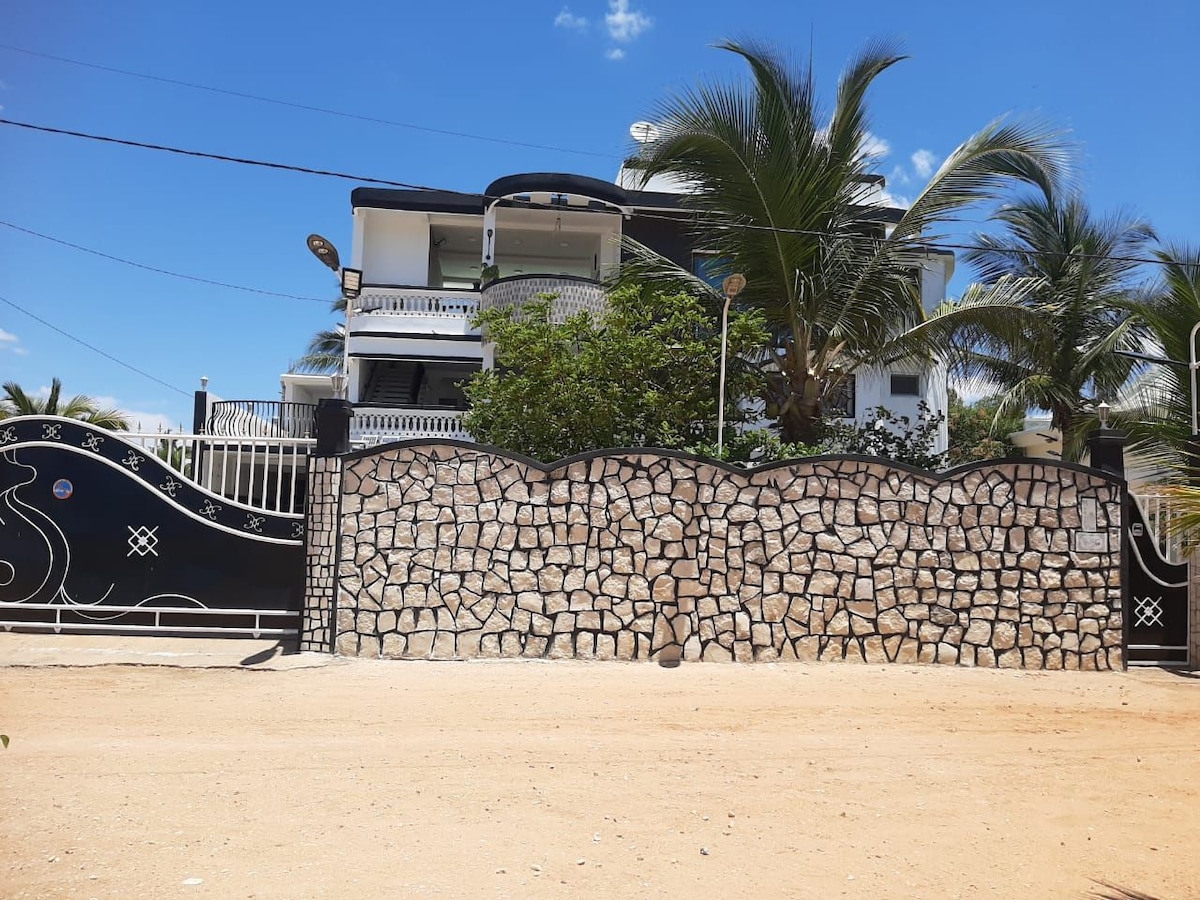 A secure property is enclosed by a stone wall and features a decorative black gate. The building above is two-storied, with a large balcony adorned with palm trees. Clear blue skies provide a bright backdrop to the scene.