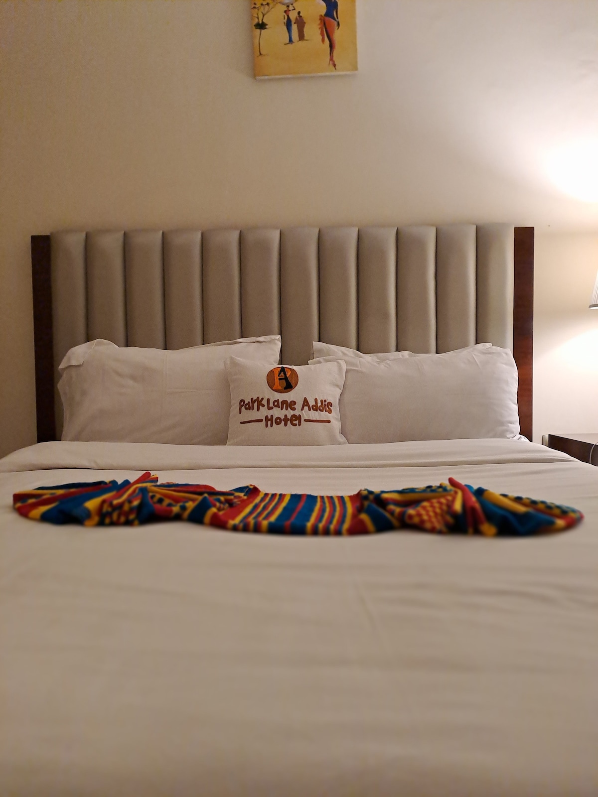 A neatly made bed is centered against a soft, padded headboard. Two plush white pillows rest against the headboard, while a colorful, patterned cloth is draped elegantly across the bedspread, adding a touch of vibrancy. A warm bedside lamp provides gentle illumination.