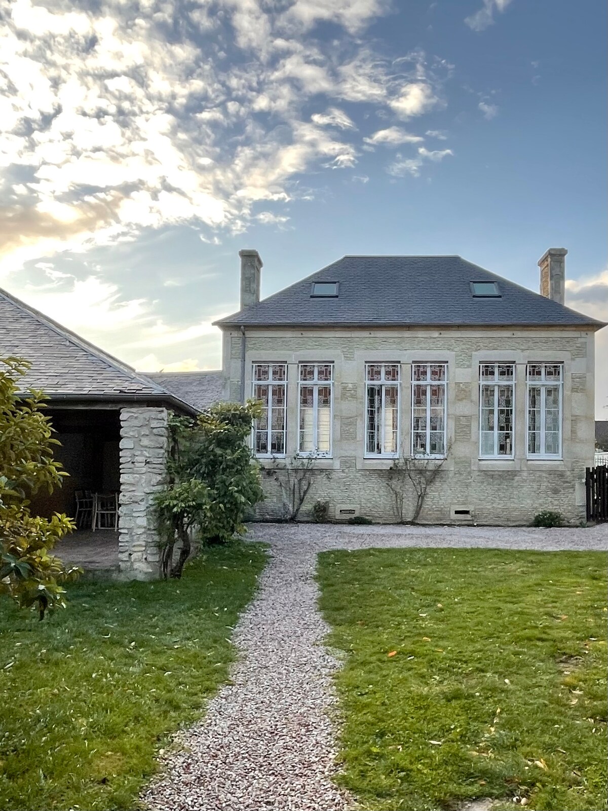 A charming stone house is depicted, featuring elegant tall windows surrounded by greenery. A gravel path leads to the entrance, bordered by a well-kept lawn. The structure showcases a pitched roof and is complemented by an adjoining covered area, adding to its inviting appearance.