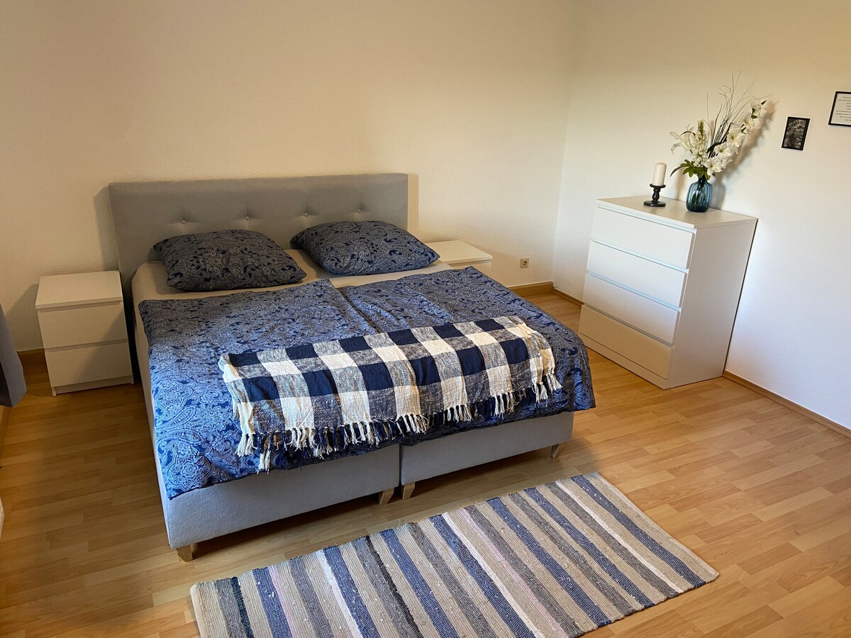 A spacious sleeping area features a comfortable bed dressed in dark blue linens and complemented by decorative pillows. A simple striped area rug adds texture to the light wooden floor. A white dresser stands nearby, accompanied by a candle and a decorative vase.