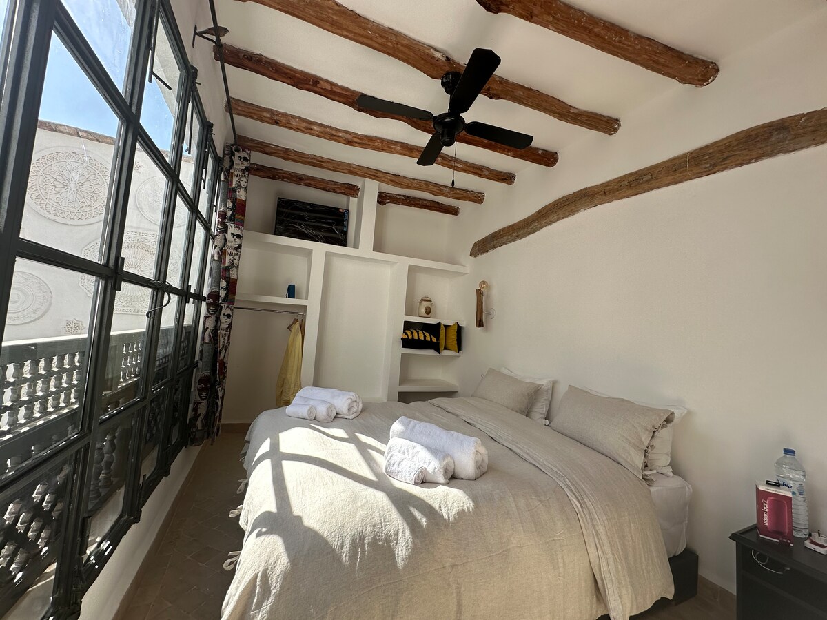 A bright sleeping area features two spacious beds with light-colored linens, positioned against a wall with exposed wooden beams. Large windows provide ample natural light, complemented by soft white curtains. A nightstand holds water and amenities.