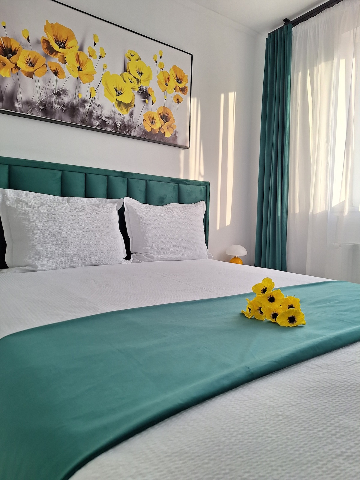 A cozy sleeping area is presented, featuring a spacious bed with white linens and a teal blanket. Yellow flowers are arranged decoratively atop the bed. Bright natural light enters through a nearby window, which is framed by soft curtain drapes.
