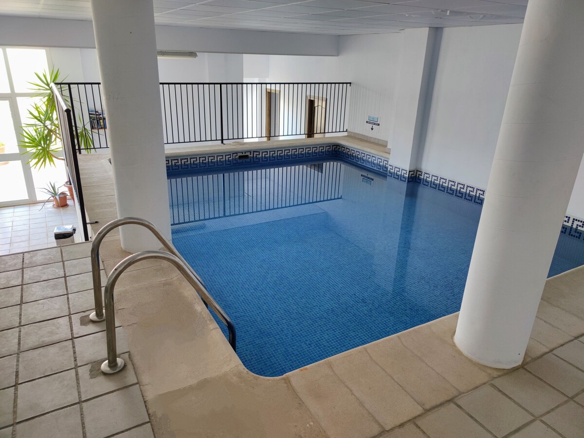 An indoor swimming pool is presented with clear blue water and a tiled border featuring a geometric pattern. A set of stainless steel steps leads into the pool. Natural light enters through nearby windows, creating a bright and inviting environment.