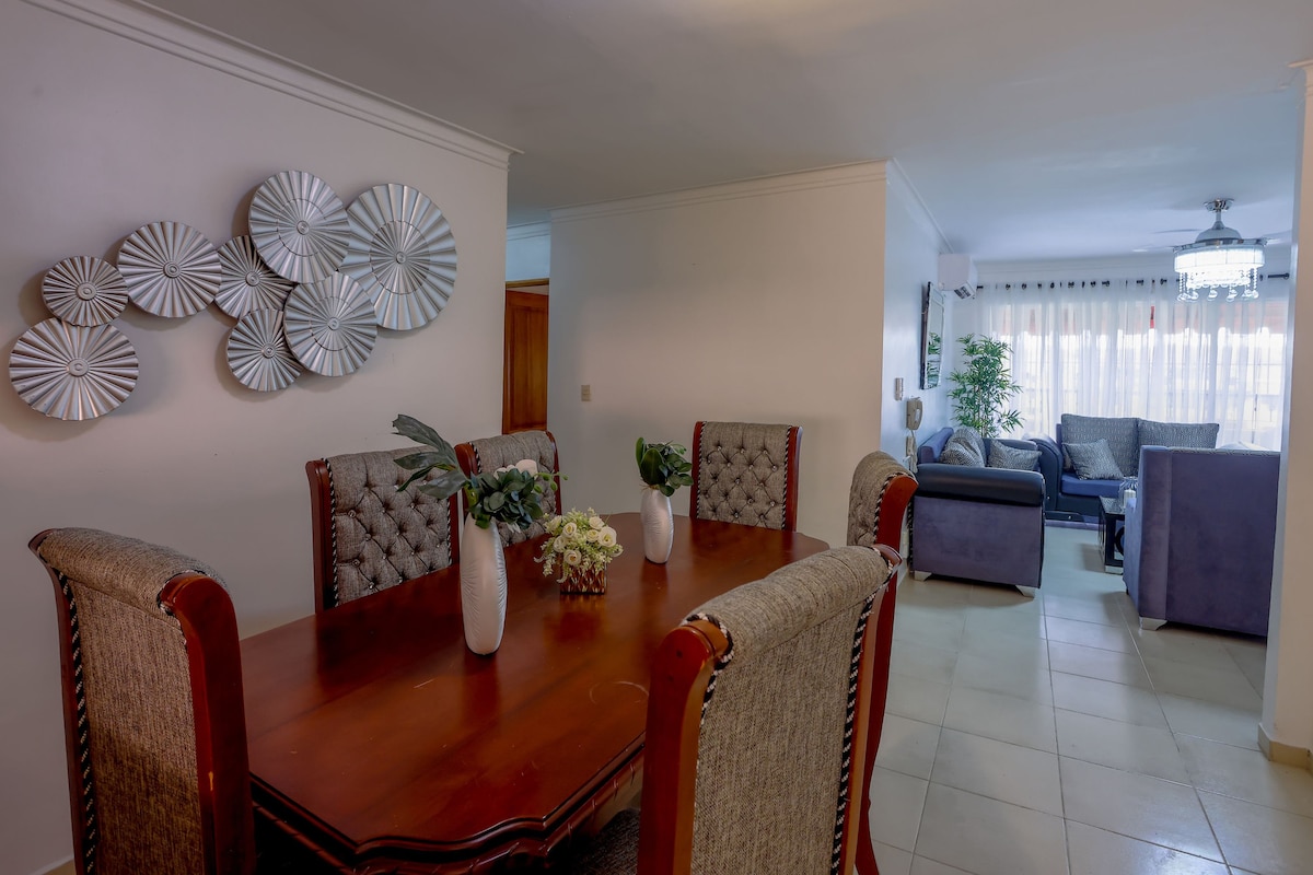 A dining area features a polished wooden table surrounded by padded chairs, complemented by a decorative arrangement of vases. The space is linked to a cozy living room, which is illuminated by natural light from large windows, offering a view of the comfortable seating.