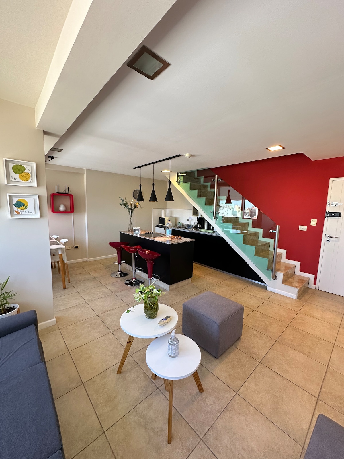 An open-plan living area showcases a modern kitchen with sleek black cabinetry and bar seating. A staircase with glass panels leads to the upper level. The space includes a comfortable couch, two small tables, and decorative items on the walls, creating a welcoming atmosphere.