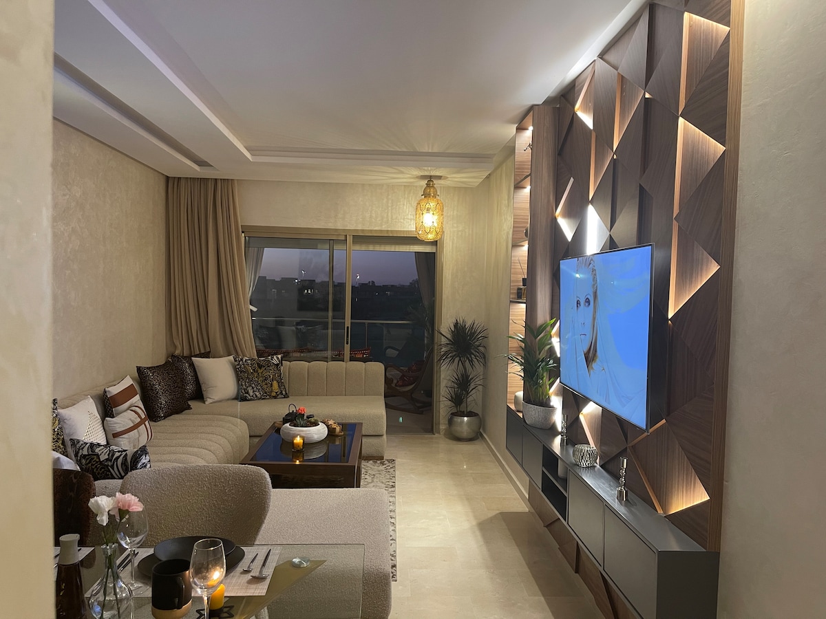 A modern living area features a large sectional sofa and a plush armchair, set beside a sleek coffee table. The wall is decorated with geometric wood panels, and a flat-screen TV is mounted for entertainment. Natural light from the nearby window creates a warm ambiance.
