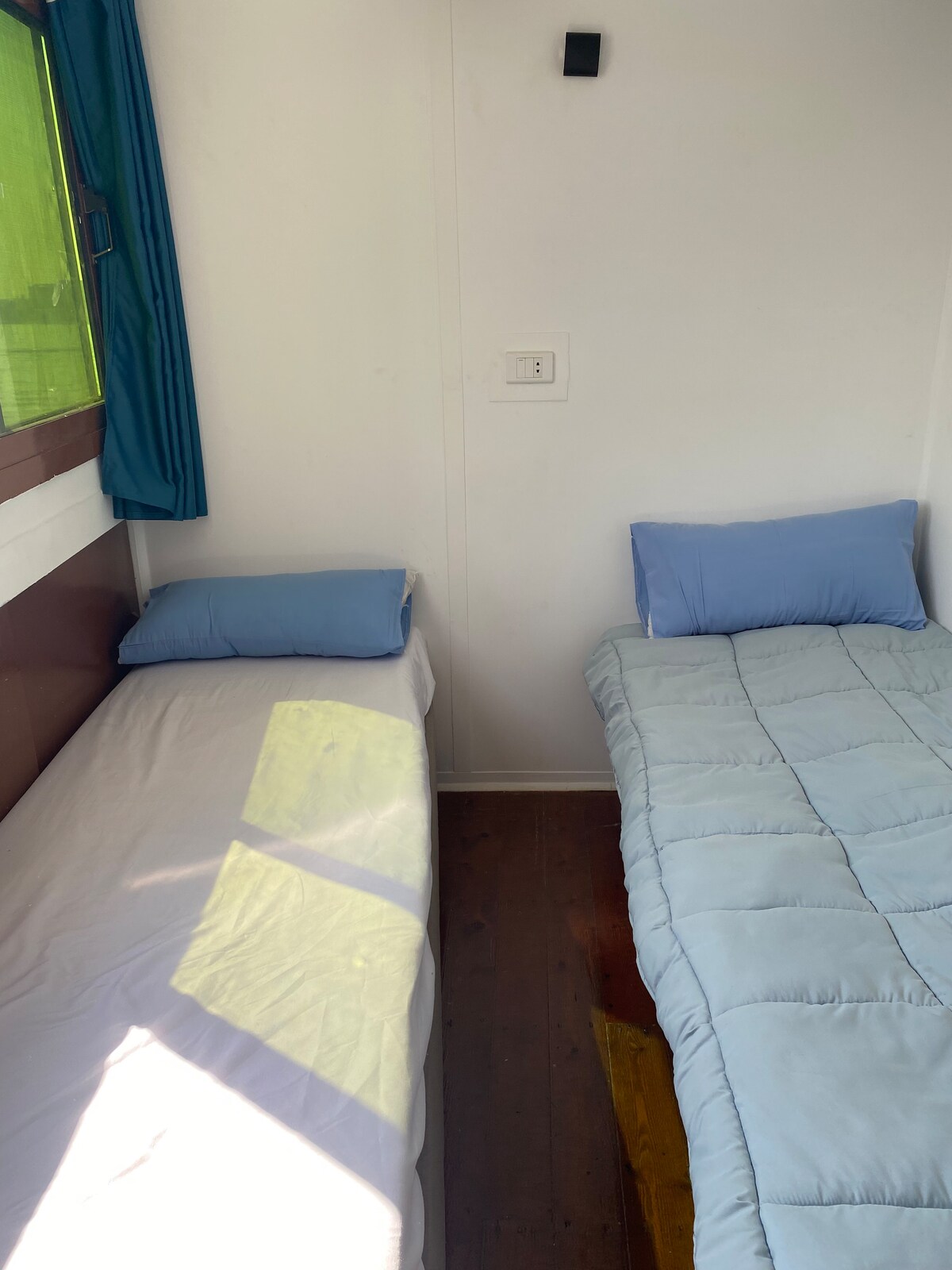 The image displays a cozy sleeping area featuring two twin beds. One bed is topped with a light blue comforter, while the other has a plain white cover. Soft blue pillows rest at the head of each bed. Natural light filters in through a window, highlighting the warm wood flooring.