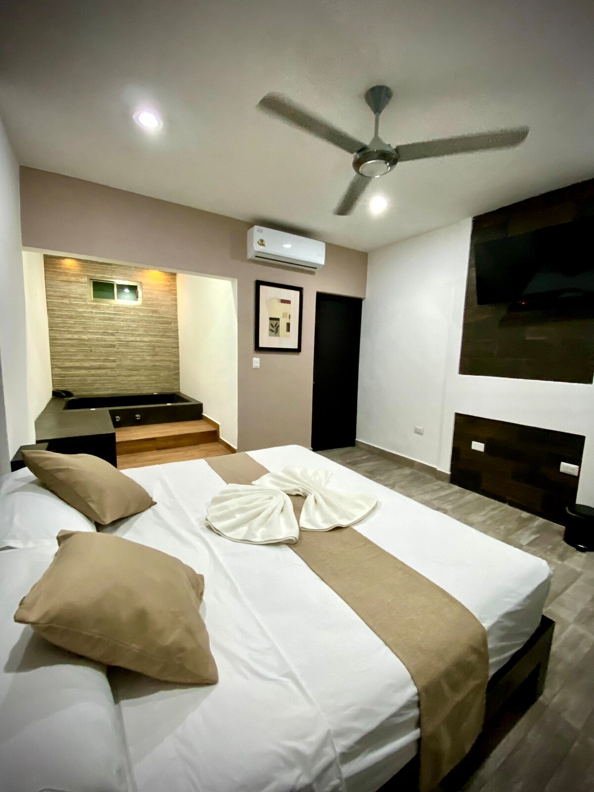 A modern bedroom features a spacious bed adorned with neutral-colored linens and decorative pillows. A ceiling fan circulates air, while a flat-screen TV is mounted on the wall. Natural light filters in from a small window, and steps lead to a separate area in the background.