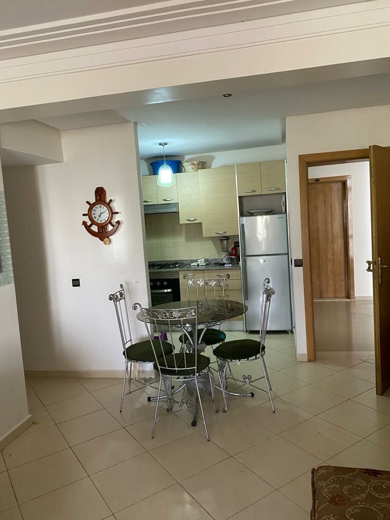 An open kitchen area is visible, featuring light-colored cabinetry and modern appliances. A round dining table with four green chairs is positioned nearby. Soft natural light fills the space, highlighting the tiled floor and a wall-mounted clock adds a nautical touch.