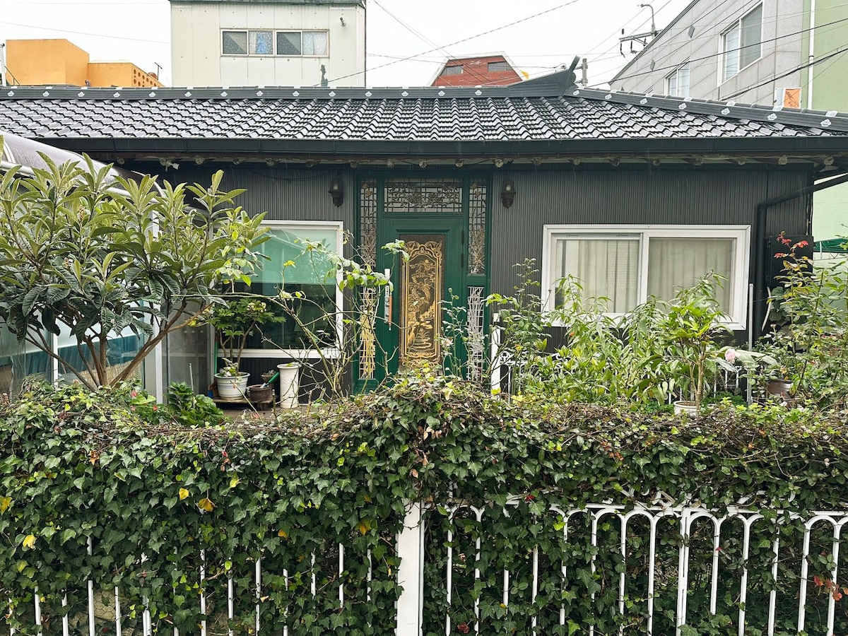 A charming exterior of a cottage is framed by a lush green hedge, with various plants visible on the porch. A decorative entrance door is set between two large windows, allowing natural light to enter. The roof features traditional tiles, contributing to the home's character.