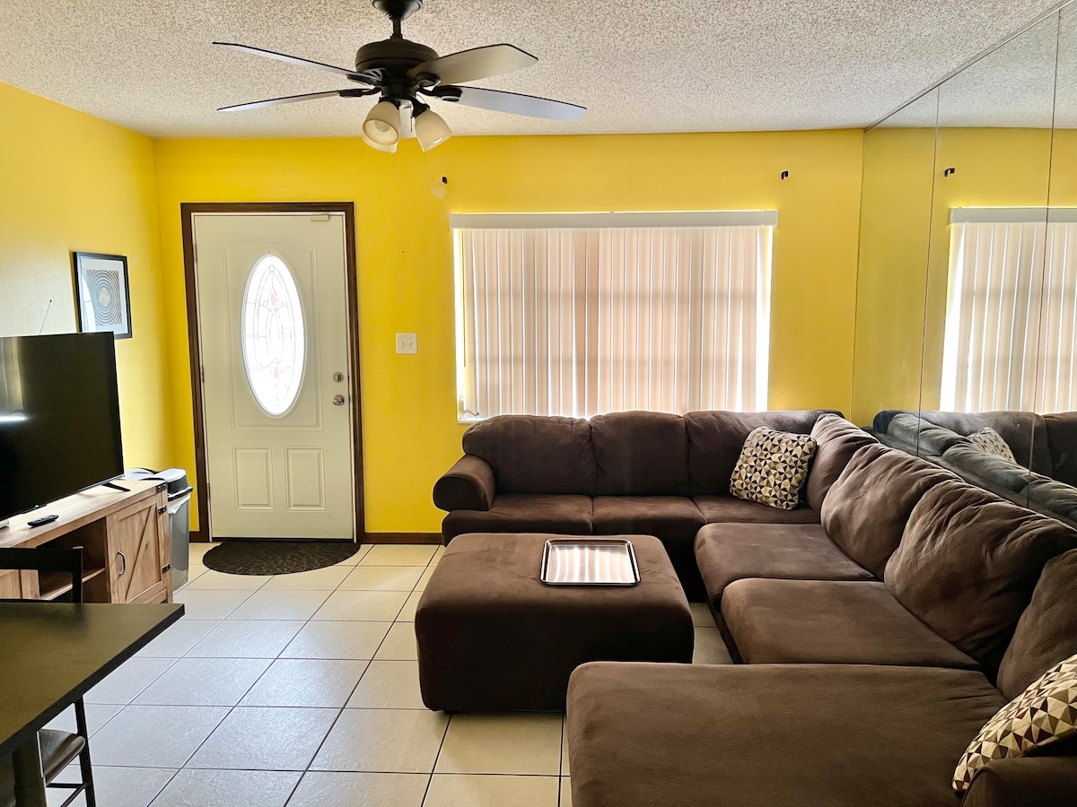 A bright living area is presented with yellow walls and a large sectional sofa. A glass coffee table sits atop tiled flooring, and a television is visible near a front door. Natural light filters through vertical blinds on spacious windows.