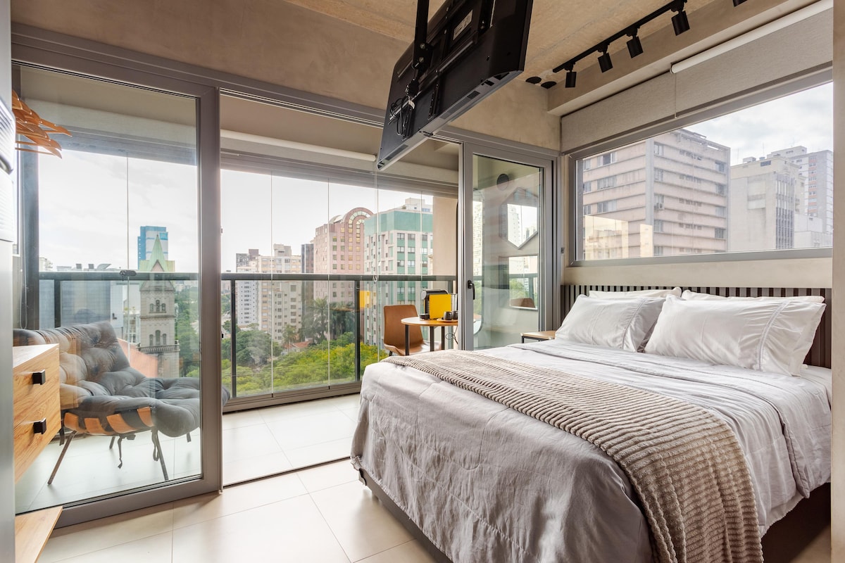 A spacious bedroom features a large bed dressed in crisp linens. Floor-to-ceiling windows allow natural light and offer views of the city skyline. A modern TV is mounted above the bed, and a small workspace is visible in the corner.
