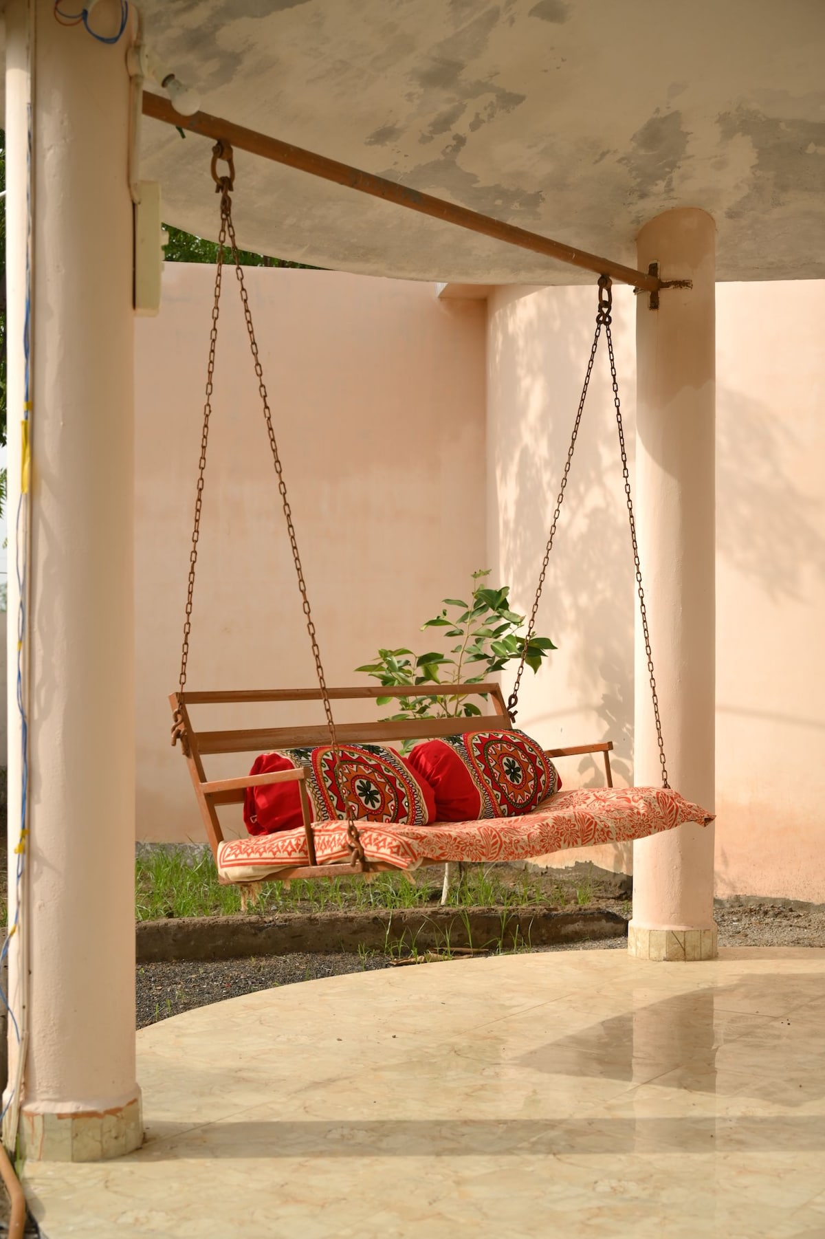 A wooden swing seat is suspended from a ceiling, adorned with vibrant, patterned cushions. The swing is positioned against a softly colored wall, and greenery is visible in the background, offering a serene outdoor space.