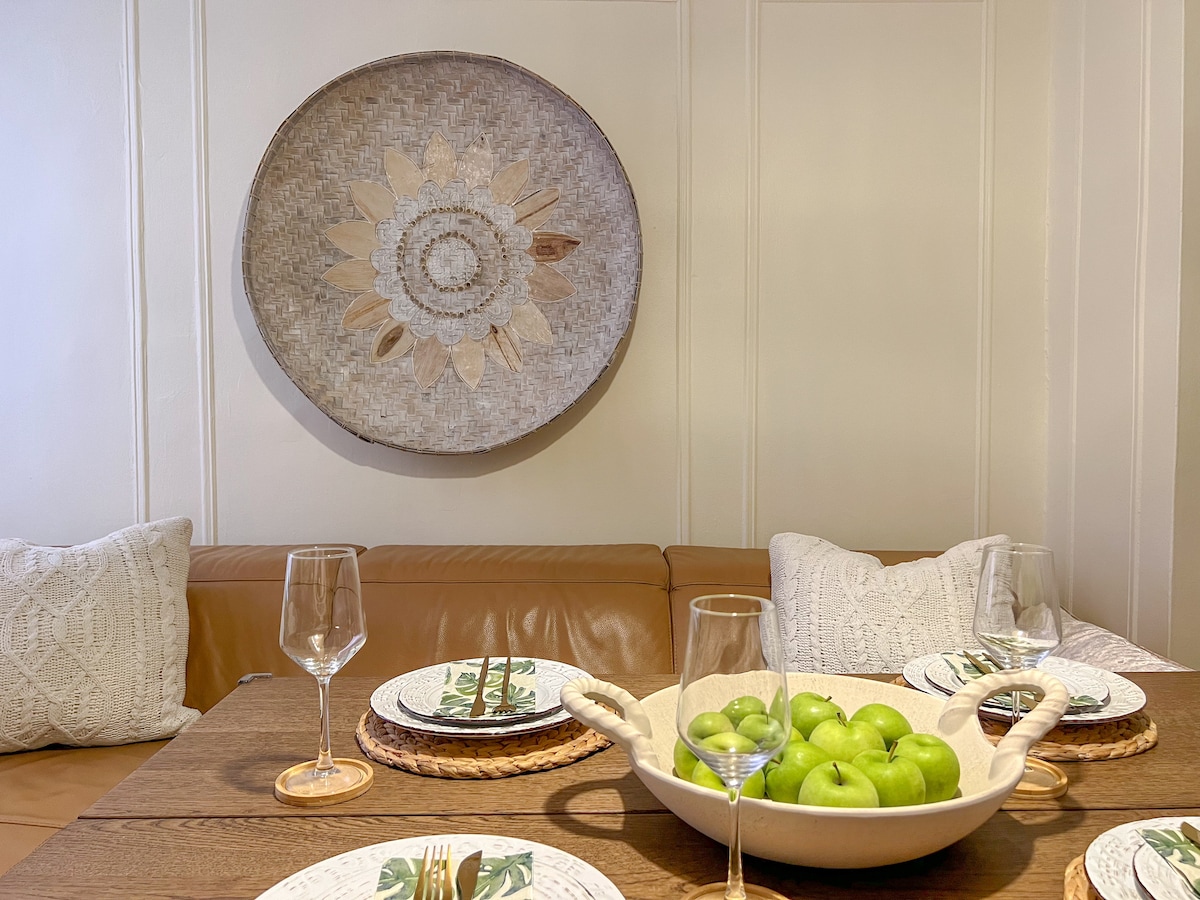 A dining area features a wooden table set with elegant glassware and decorative plates. A large bowl of green apples is centered on the table, while cushions adorned with intricate designs are arranged nearby. A round, woven art piece is mounted on the wall.