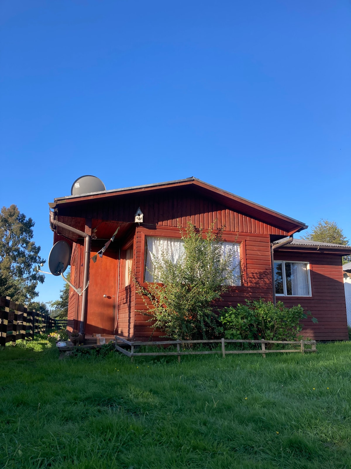 The exterior of a rustic wooden cabin is visible, surrounded by lush green grass. Satellite dishes are mounted on the roof, and a wooden fence borders the property. Clear blue skies create a peaceful backdrop for the home.