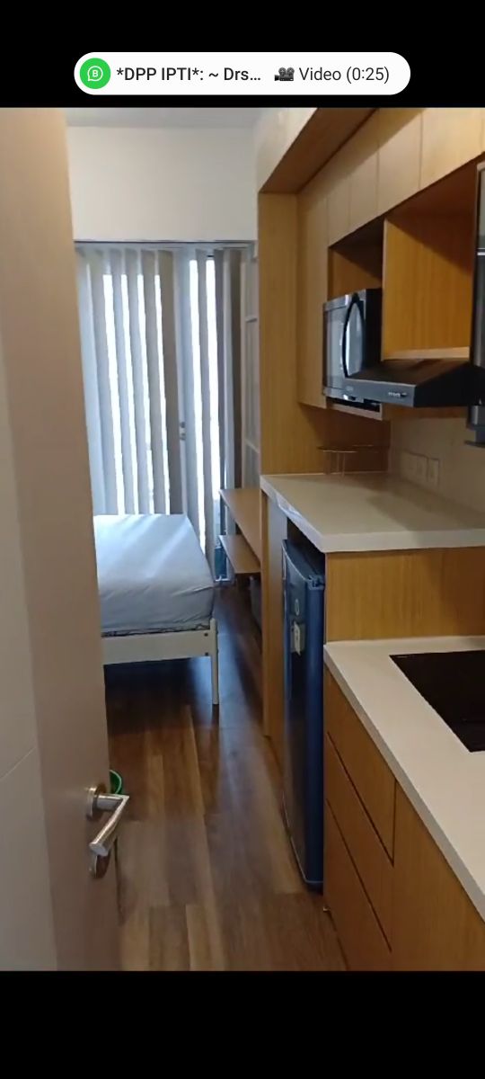 A streamlined kitchen area is highlighted by warm wooden cabinetry and modern appliances. A compact dining space is visible beside the kitchen. Natural light filters through vertical blinds onto a minimalistic sleeping area furnished with a ready bed, enhancing a sense of openness.