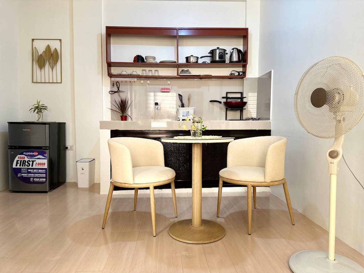 A dining area is presented with a round table and two cushioned chairs. A compact kitchen can be seen in the background, featuring a range of appliances and cabinetry. A small refrigerator is positioned nearby, and a fan stands to the side, providing additional comfort.