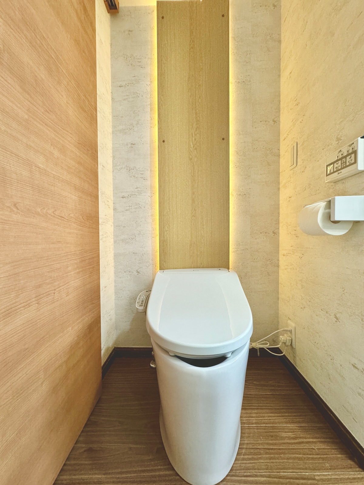 The image features a clean, minimalist bathroom with a modern toilet. Soft, indirect lighting highlights the wooden paneling behind the toilet. The floor is wooden, and a toilet paper holder is mounted on the wall nearby, creating a functional and simple space.