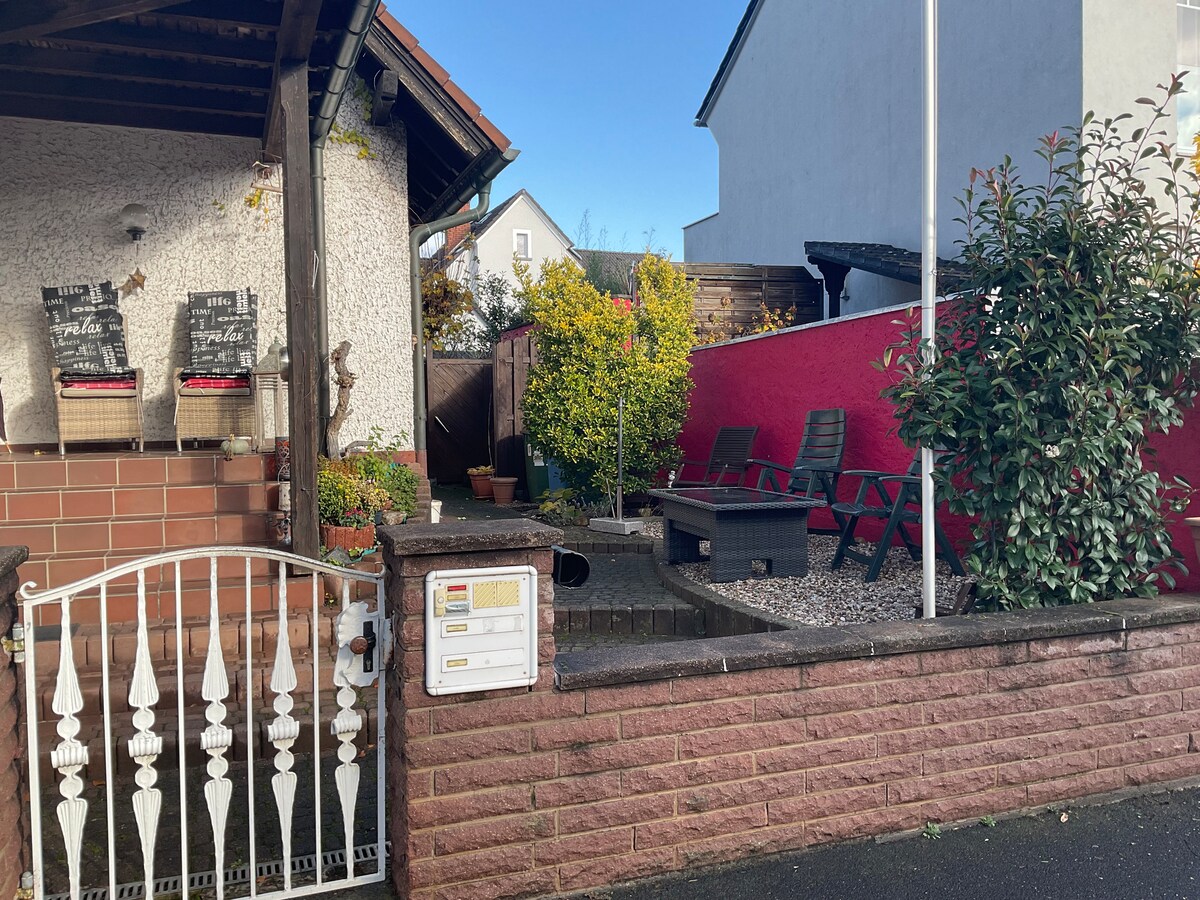 A quaint entrance is seen with a gated pathway leading to a landscaped area. Comfortable outdoor seating in a secluded corner is framed by decorative plants and a vibrant red wall, offering a cozy spot for relaxation. The exterior showcases a blend of textures and inviting elements.