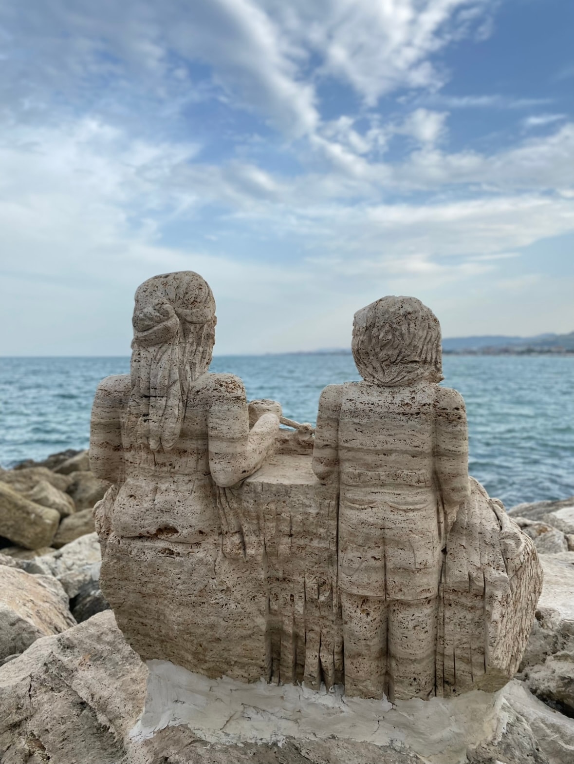 A stone sculpture depicts two figures seated side by side, overlooking the calm sea. The textured surface of the stone contrasts with the smooth water, while soft clouds drift in the blue sky above, creating a serene coastal scene.