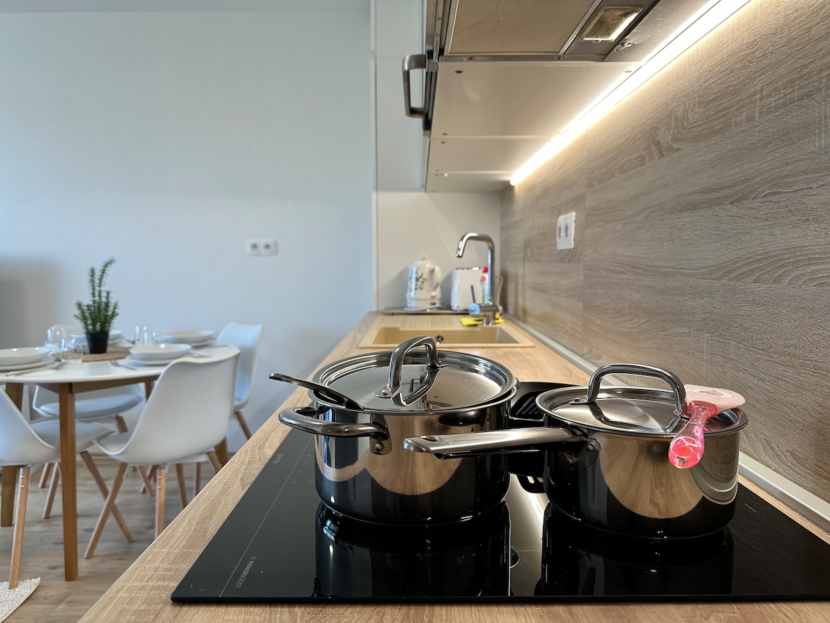 A well-equipped kitchen showcases a sleek stovetop with two pots, the handles glinting in the light. In the background, a dining table is set for a meal, surrounded by four white chairs against a minimalist design.