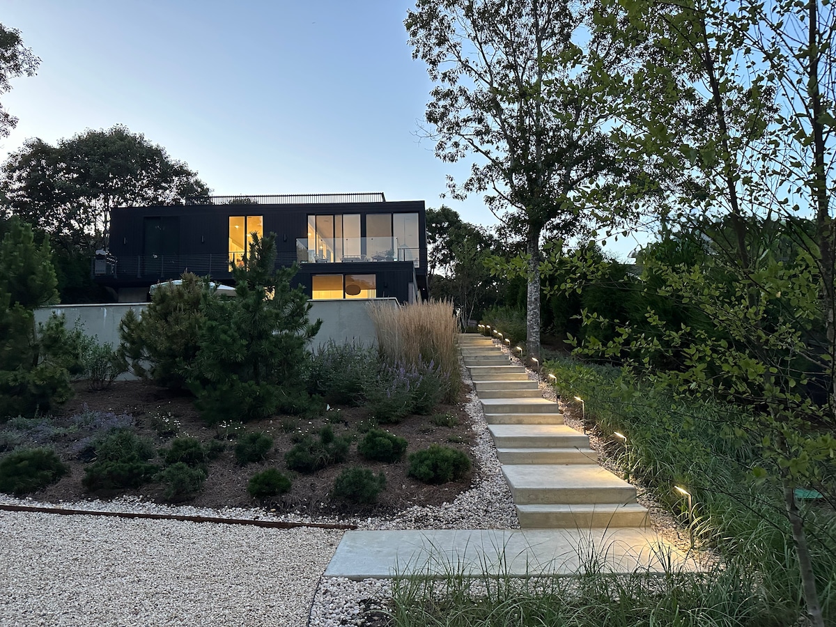 A modern home is set against a twilight sky, accessible via a well-lit stone pathway lined with greenery. The landscaped garden features low shrubs and ornamental grasses, while the house is designed with large windows that provide a glimpse of the inviting interior.