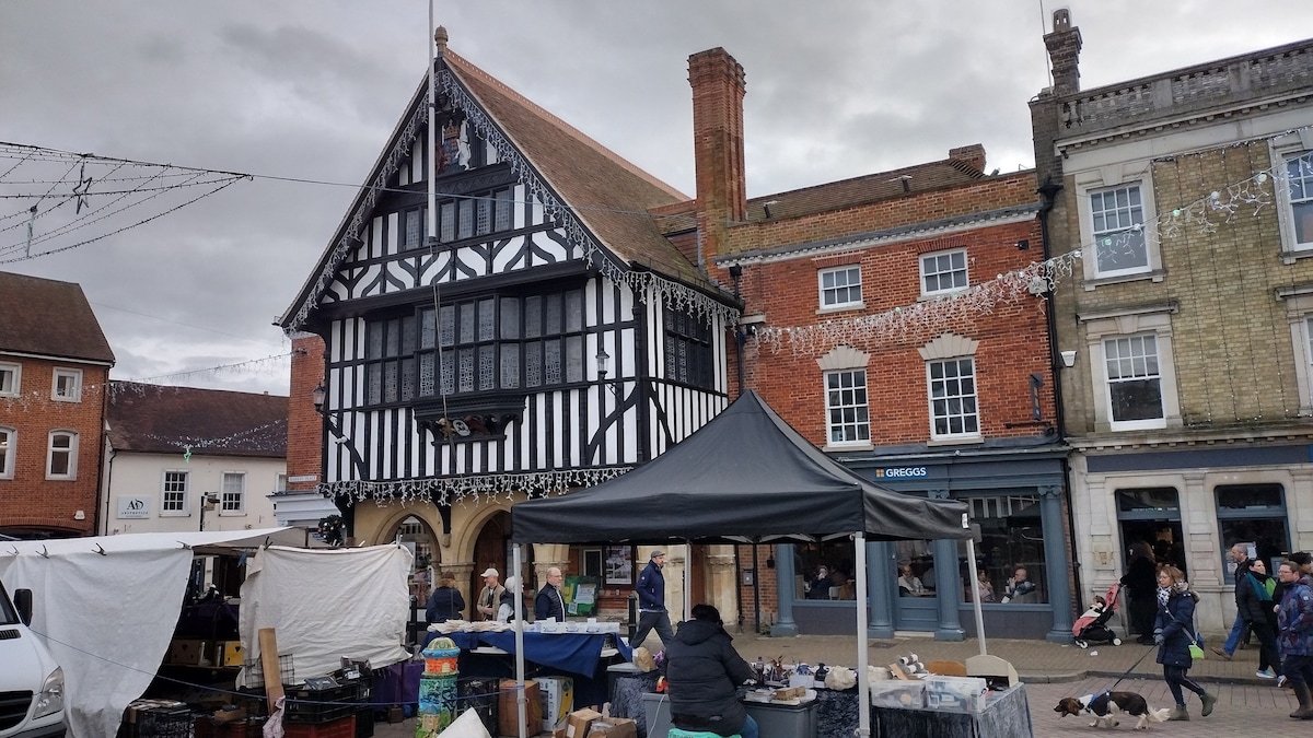 A historic building with black and white timber framing is set in the center of a lively market square. Stalls with a variety of goods are arranged in the foreground, while pedestrians can be seen strolling by amidst festive decorations hanging overhead.