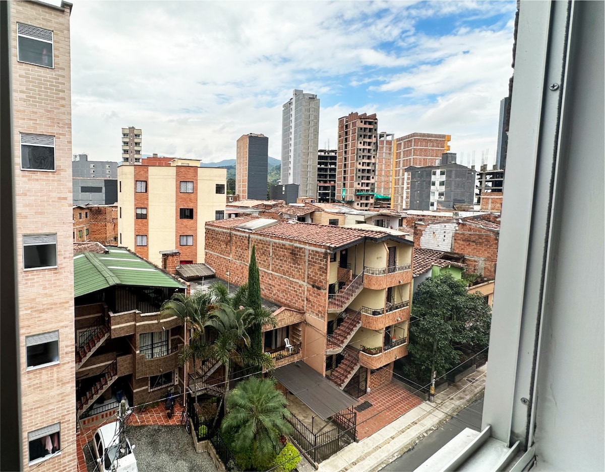 A view from the window displays a mix of modern and traditional architecture. Nearby buildings feature multiple stories, with brick facades and greenery interspersed. The skyline is highlighted by a backdrop of taller structures under a partly cloudy sky.