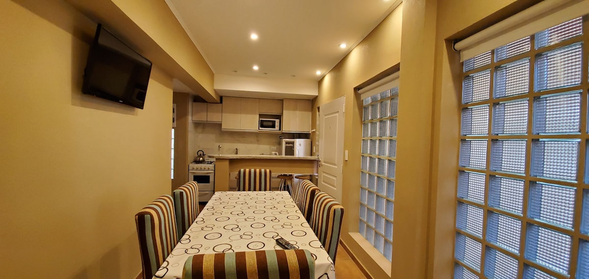 An open dining and kitchen area is presented, featuring a long table surrounded by striped chairs. A television is mounted on the wall, and modern kitchen appliances are visible. Glass block windows allow natural light to enter, enhancing the overall bright atmosphere.