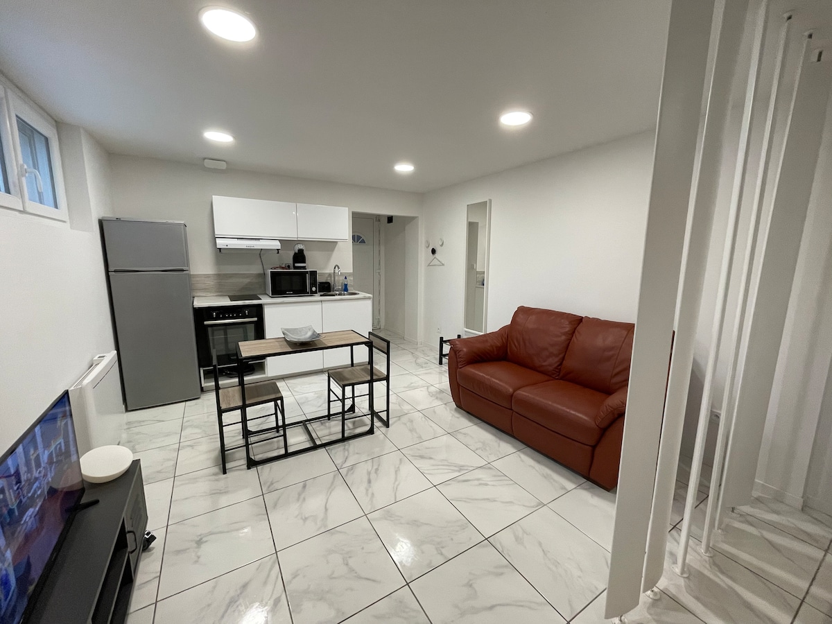A bright and well-equipped living space features a leather sofa, a coffee table, and a television. The kitchen area is fitted with modern appliances, including a refrigerator and an oven. White walls and marble tile flooring contribute to a clean and open atmosphere.
