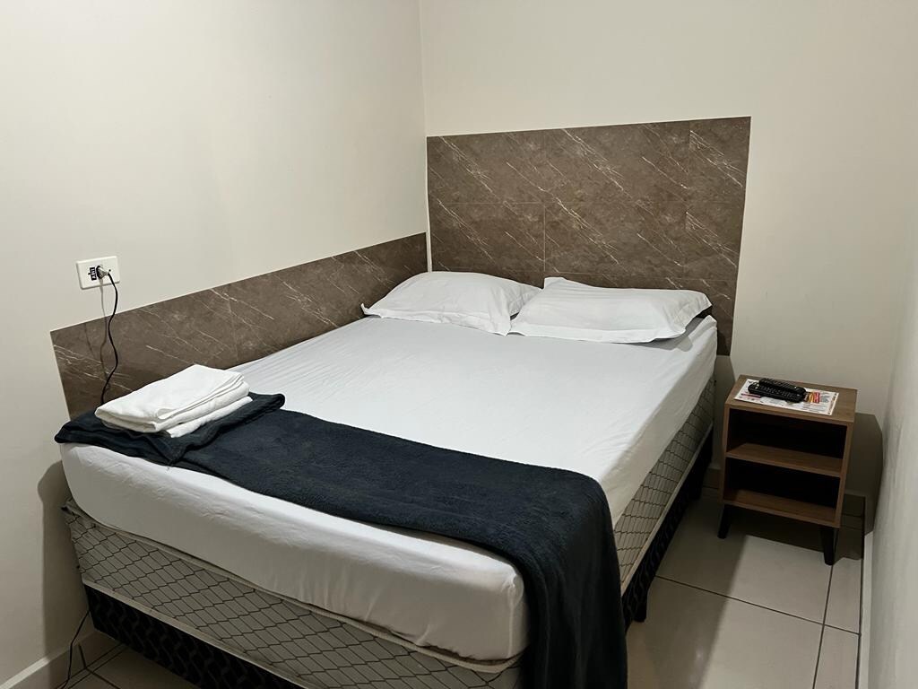 A cozy bedroom showcases a spacious bed with a white mattress and neat linens. A dark throw blanket is laid across the bed. A bedside table is positioned next to the bed, featuring a small stack of magazines and a remote control.