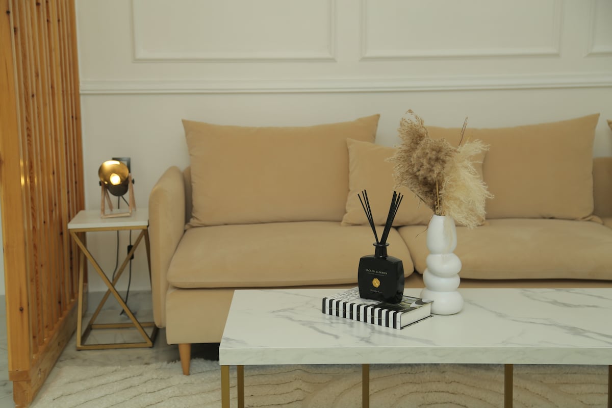 A light-colored couch is positioned against a smooth wall, complemented by a marble-topped coffee table. A decorative vase with dried flowers sits atop the table, alongside a black decorative item and a striped book. A softly lit lamp adds warmth to the space.
