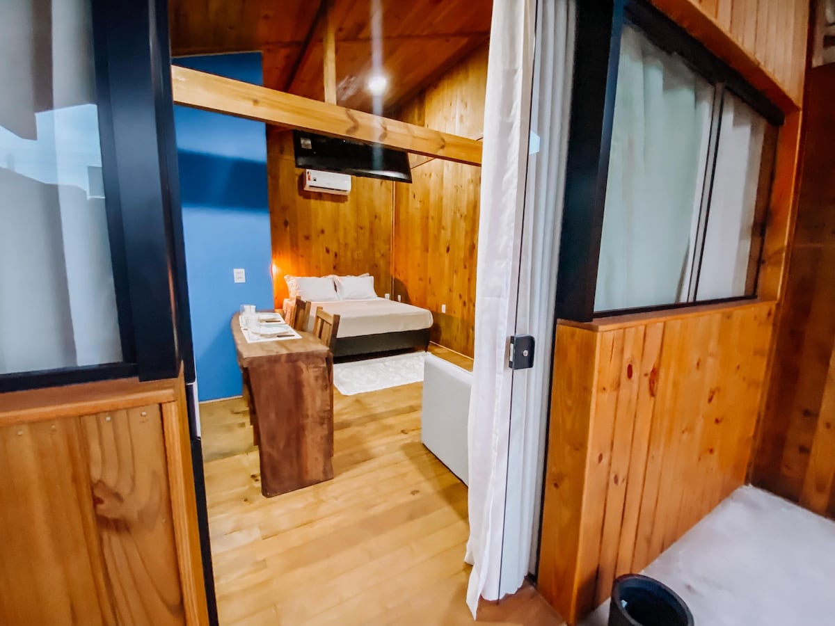 A warmly lit interior space is presented, featuring wooden walls and a cozy layout. A spacious bed is visible in the background, along with a small table and seating area. Natural light filters through large windows, enhancing the inviting atmosphere.