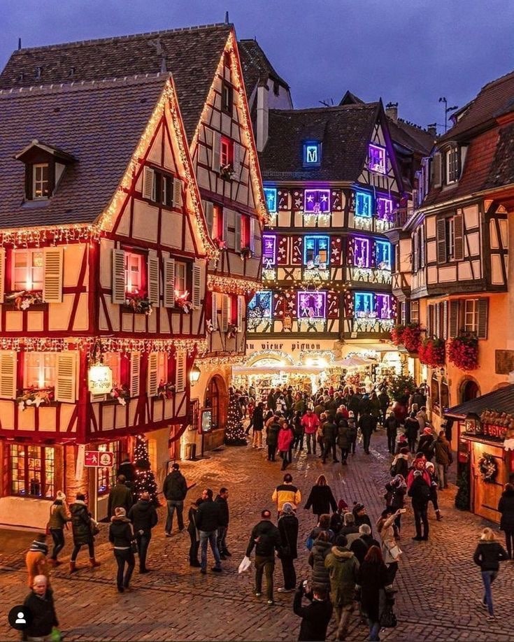 A charming street scene features traditional half-timbered buildings adorned with festive lights and decorations. Crowds of visitors stroll along the cobblestone path, while shop windows display holiday decor. The atmosphere reflects a vibrant community during the evening hours, enhanced by the warm glow of lights.