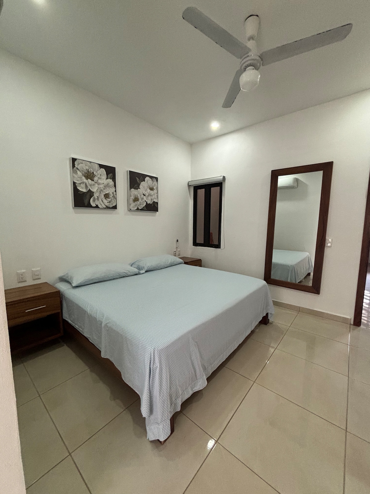 A well-lit bedroom is presented with a spacious bed dressed in light blue linens. Two floral wall art pieces adorn the white walls, and a large mirror reflects the room's features. A minimalist wooden nightstand is positioned beside the bed, and a ceiling fan offers ventilation.