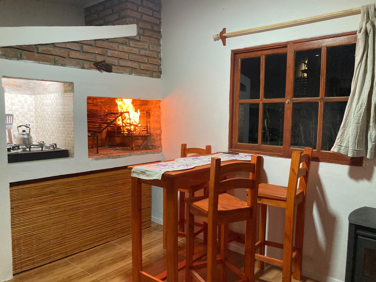 A cozy dining area features a tall wooden table with four chairs, set against a brick wall. A fireplace is visible, with flames adding warmth to the space. Natural light filters through a window adorned with light curtains, enhancing the inviting atmosphere.