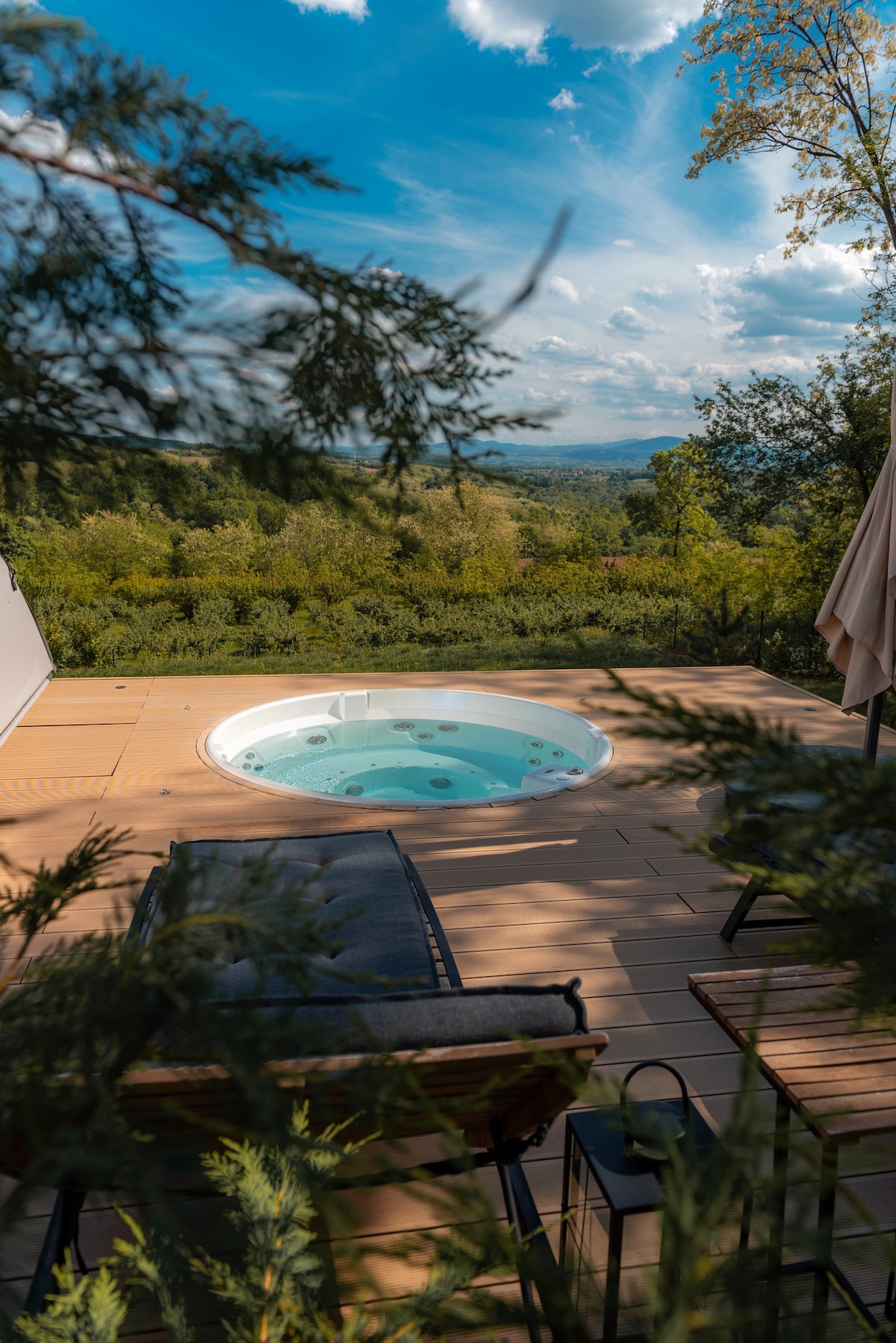 A private outdoor hot tub is nestled on a wooden deck, surrounded by lush greenery and offering expansive views of the landscape. Comfortable seating is placed nearby, inviting relaxation amidst the natural surroundings under a clear blue sky.