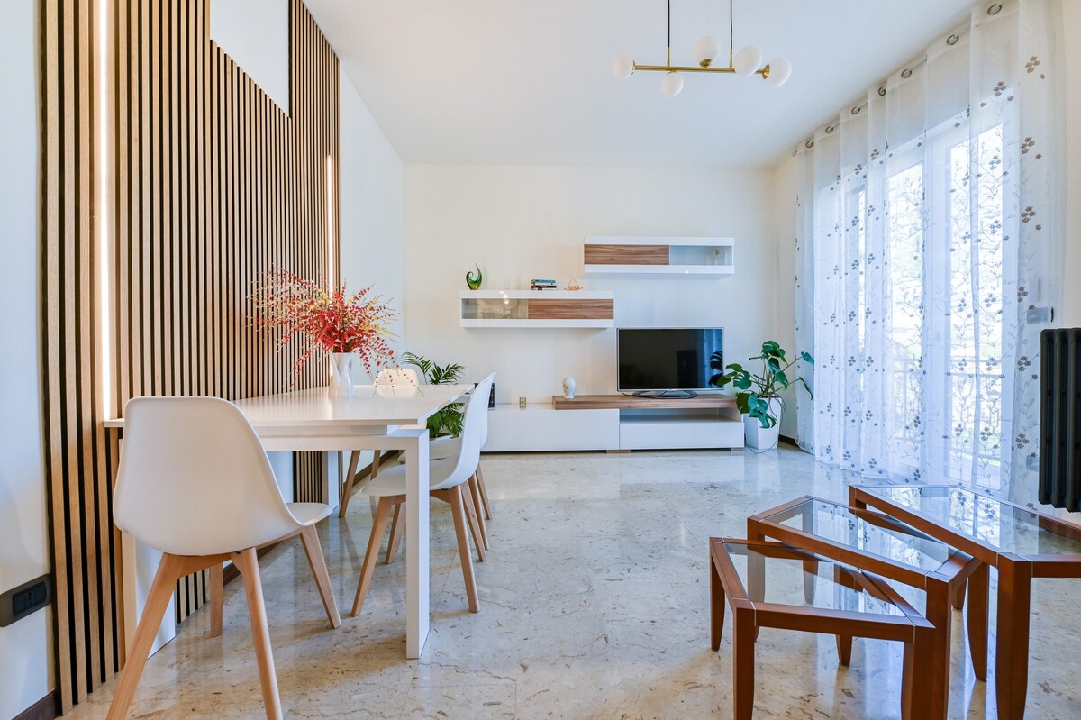 A modern living area features a light-colored tiled floor and wooden accent walls. A white dining table with four chairs is positioned toward the front, accompanied by a sleek entertainment unit with a television. Large windows, adorned with sheer curtains, allow natural light to fill the space.