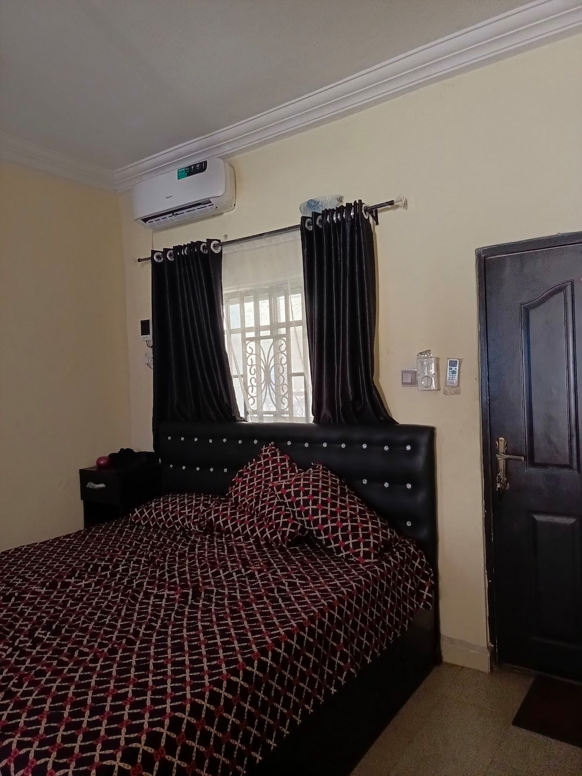 A cozy bedroom features a spacious bed dressed in a patterned duvet. Black curtains frame the window, allowing natural light to enter. An air-conditioning unit is mounted above, and a dark wood door leads to another area of the property.