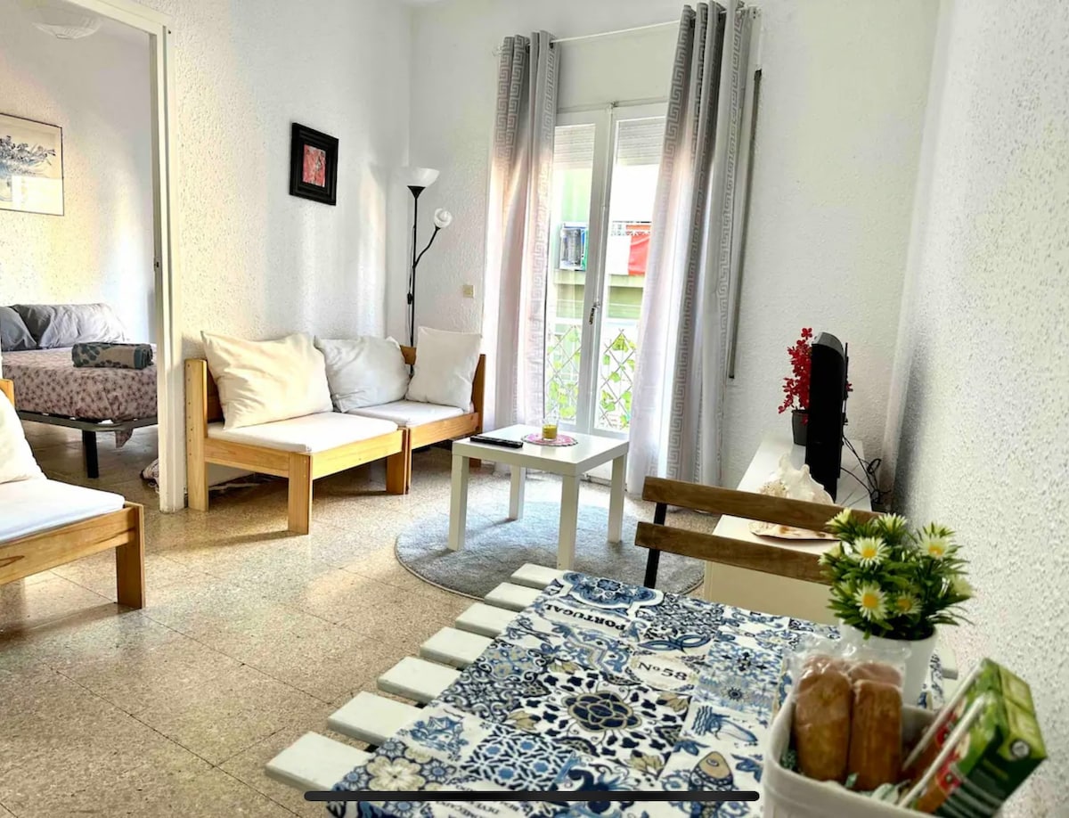 A bright living area features two light-colored sofas arranged around a small, round table adorned with flowers. A lamp stands in one corner, and large windows with sheer curtains provide natural light, showcasing a view of the outside greenery.