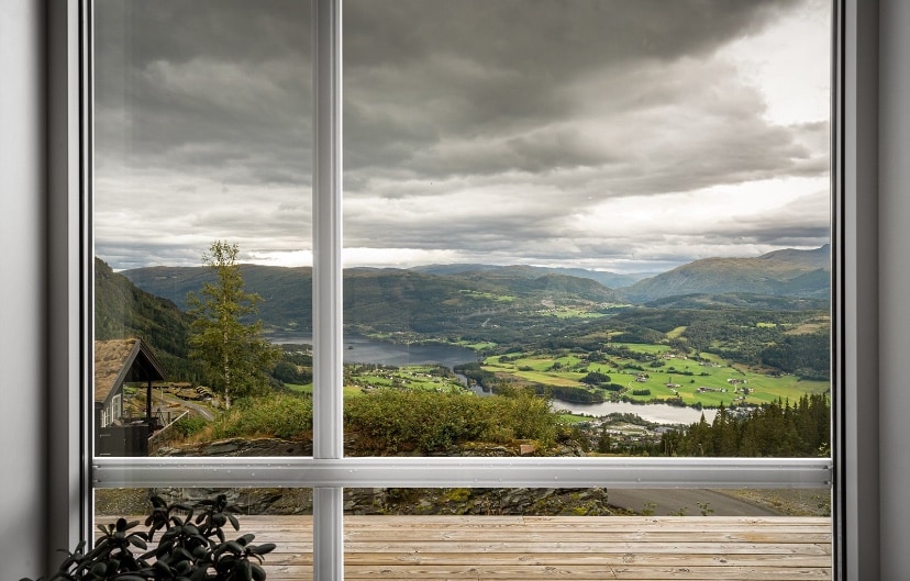 A large window frames a panoramic view of rolling hills and a serene lake. Overcast skies are visible, with a hint of greenery from surrounding trees and fields. Natural light floods the interior space, enhancing the scenic backdrop.