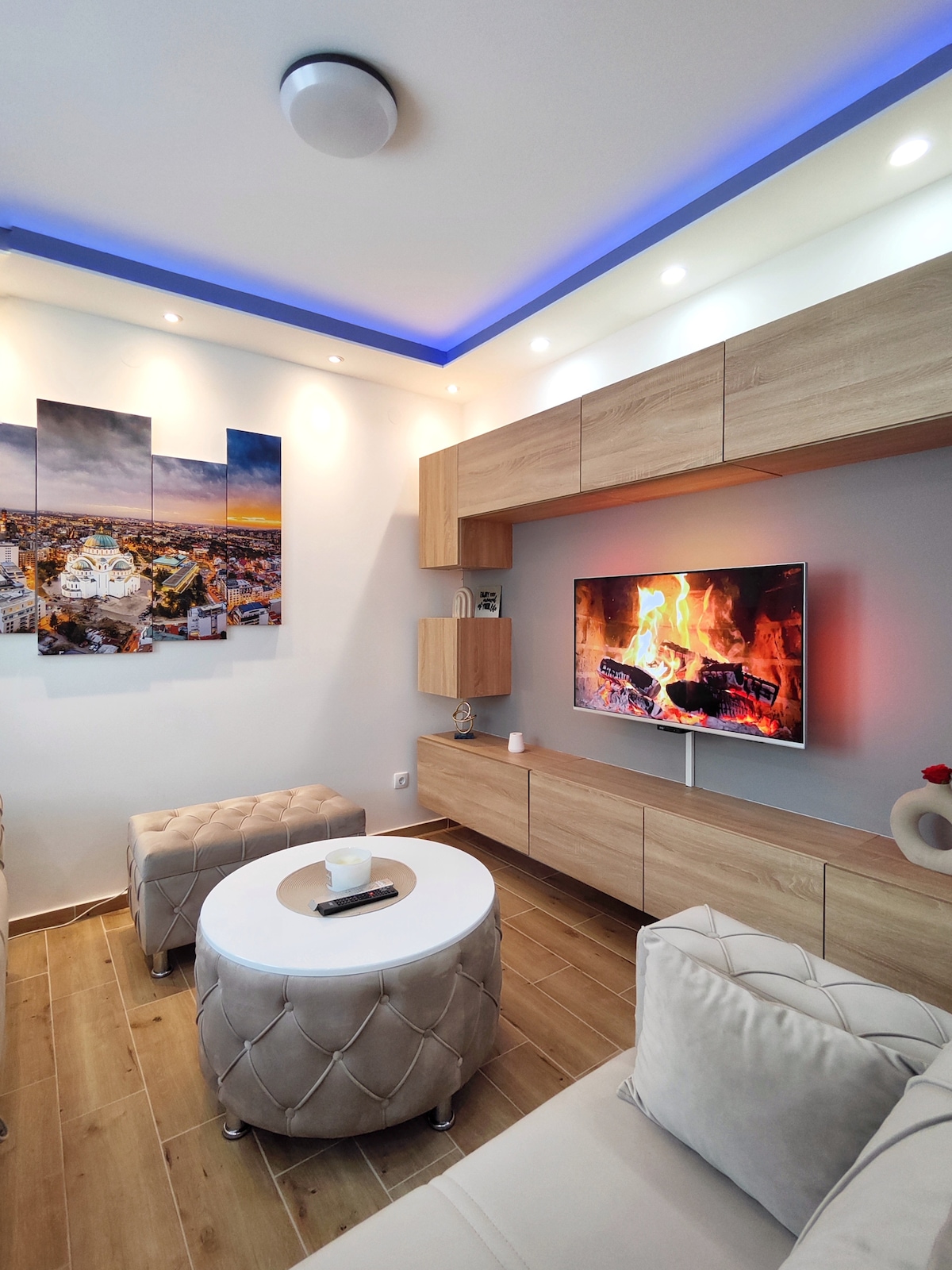 A modern living area is furnished with light-colored, plush seating arranged around a circular coffee table. A wall-mounted television displays a cozy fireplace scene. Soft blue lighting highlights the ceiling, while an artwork depicting a cityscape is featured on one wall.