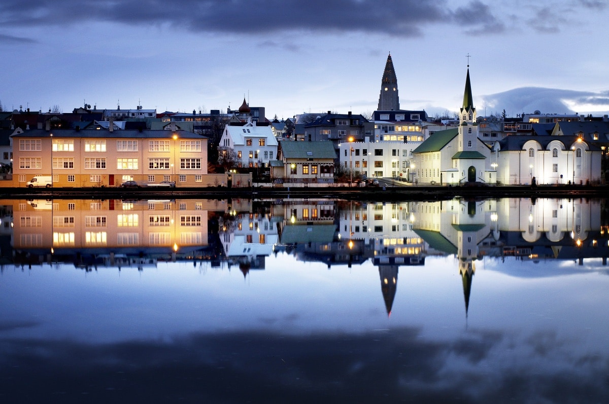 A serene urban landscape is reflected in calm waters, featuring a mix of illuminated buildings and a prominent church steeple. The scene is set against a twilight sky, highlighting the architectural details and vibrant colors of the structures along the waterfront.