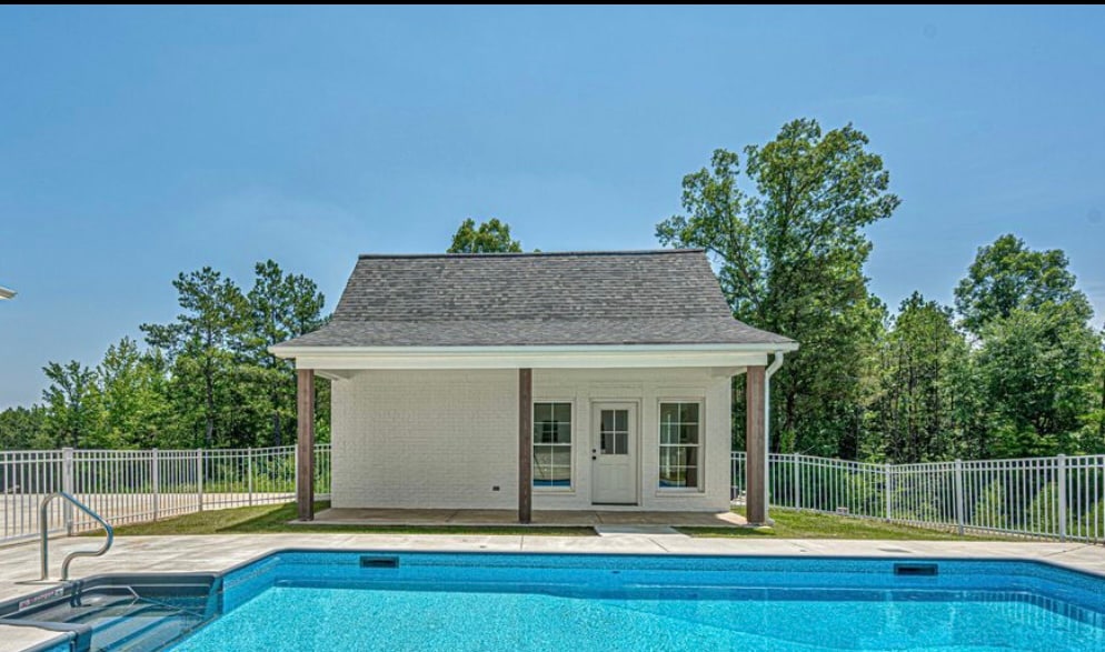 A serene swimming pool is surrounded by a spacious patio, featuring a cozy structure with large glass doors. Lush greenery frames the scene, providing a natural backdrop. The blue water of the pool reflects the clear sky above.