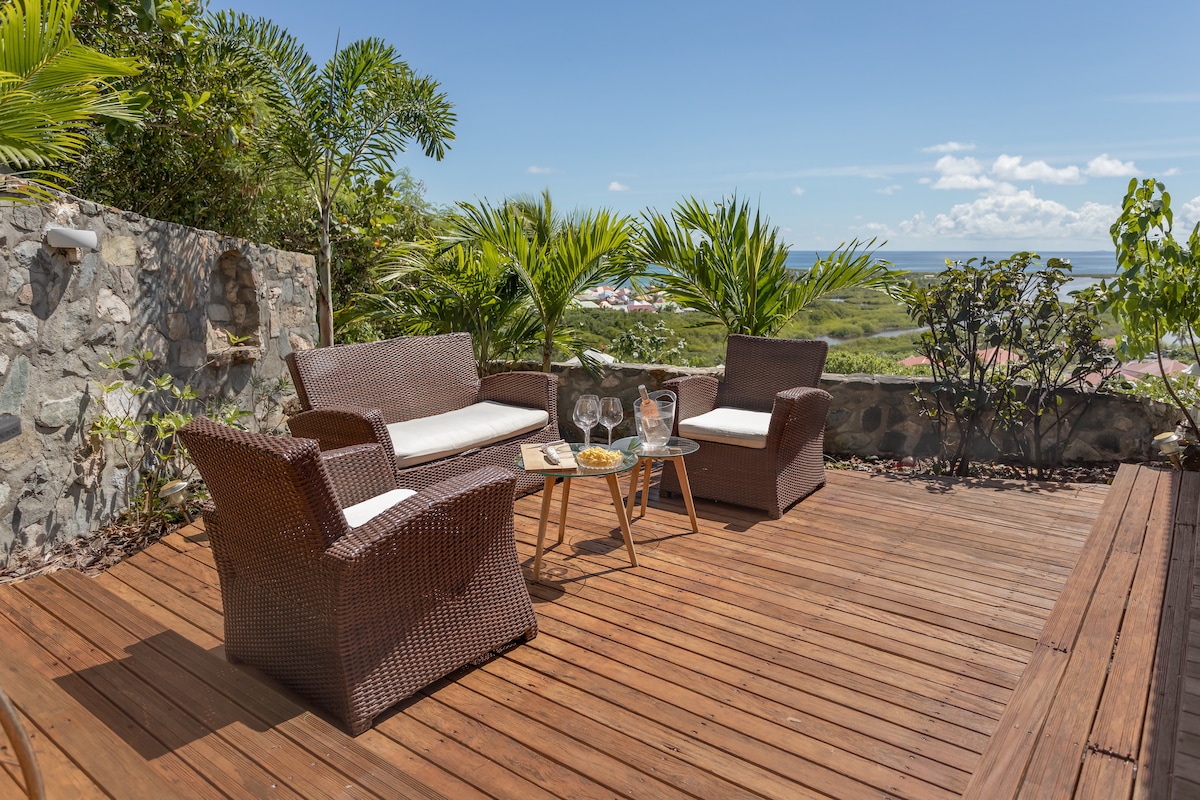 An outdoor terrace is furnished with two wicker chairs and a small round table. Glassware is neatly arranged on the table, surrounded by lush greenery and stone accents. A scenic view of the landscape is visible in the background, under a clear blue sky.