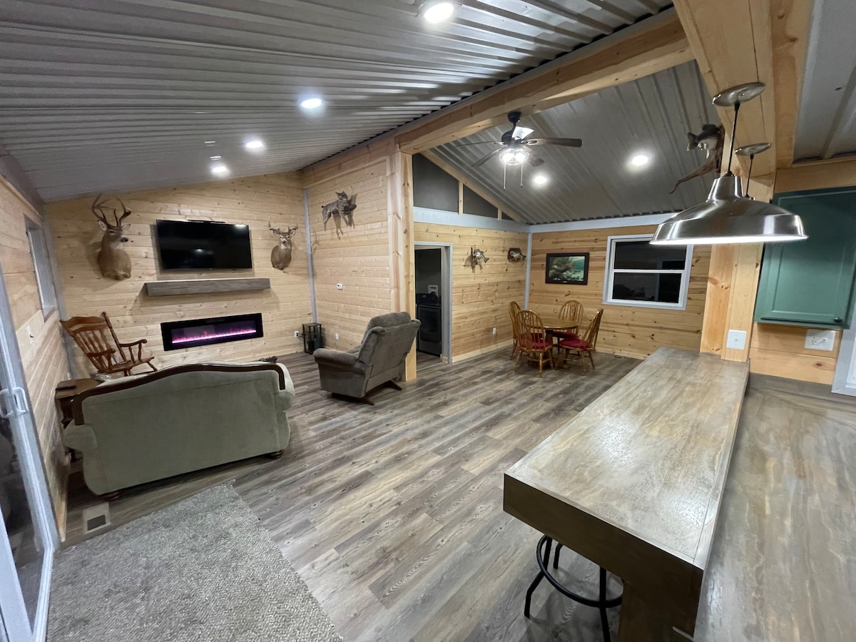 A spacious living area features wooden paneling and a modern electric fireplace. Comfortable seating options include a recliner and a sofa. A dining table is visible in the corner, surrounded by chairs. Natural light fills the space through large windows, enhancing the open feel.