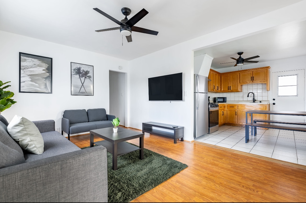 The living area features a comfortable gray sofa and a matching armchair, complemented by a coffee table on a soft rug. A wall-mounted TV is positioned above a low media unit, while a spacious kitchen with wooden cabinets is visible in the background.