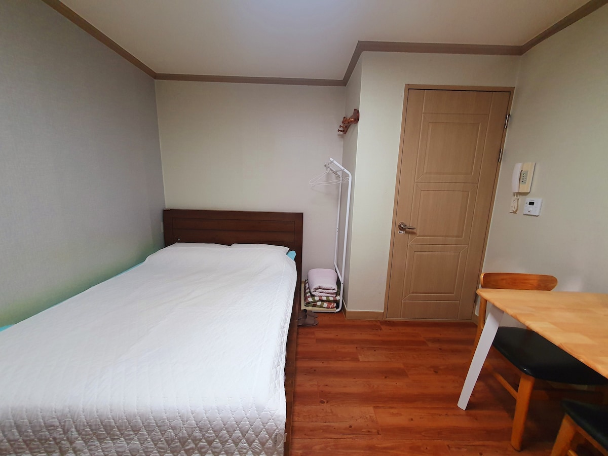 A simple room is presented, featuring a spacious bed with a light-colored coverlet. A wooden table and chair are positioned near the wall, accompanied by a clothing rack. The warm-toned flooring complements the muted wall colors, contributing to an inviting ambiance.